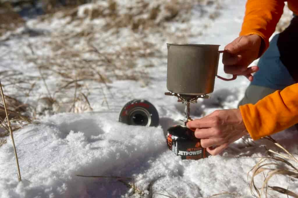 A close up of a hiker holding the evernew pasta pot on a stove with one hand and turning the stove on with the other. The lid of the pot is sticking out of snow.