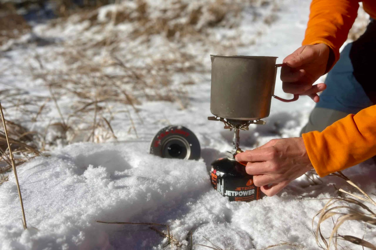 A close up of a hiker holding the evernew pasta pot on a stove with one hand and turning the stove on with the other. The lid of the pot is sticking out of snow.