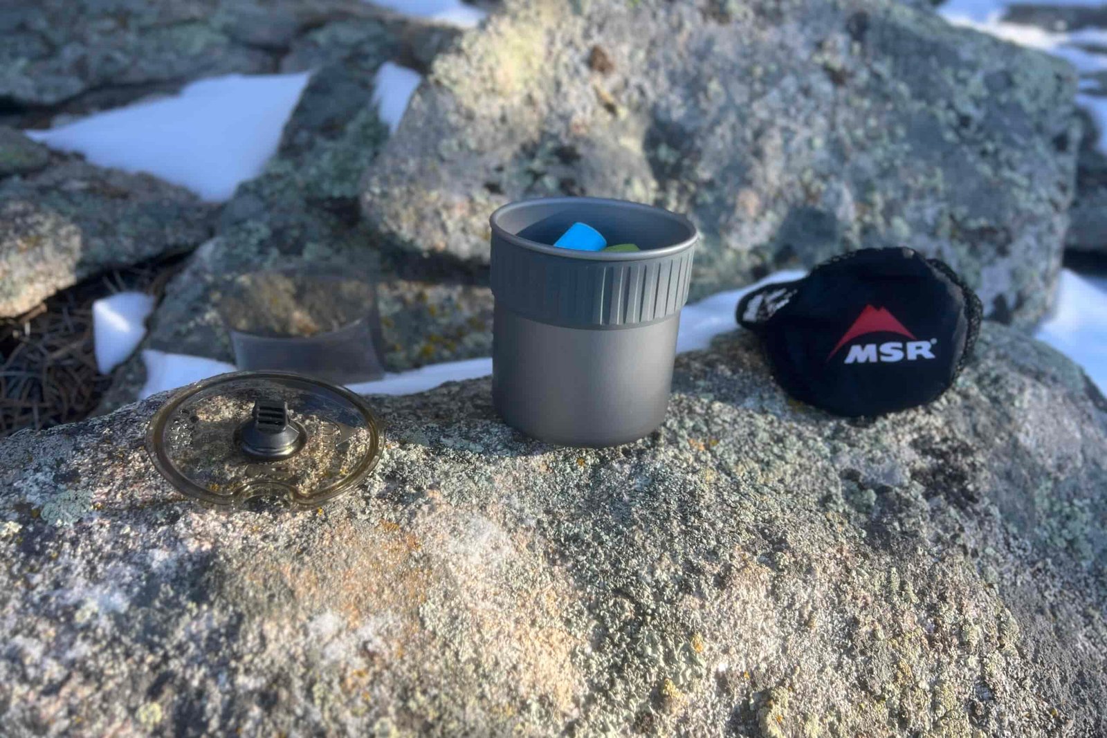 The lid, pot, and stuff sack of the msr trail mini solo sit next to each other on a rock.