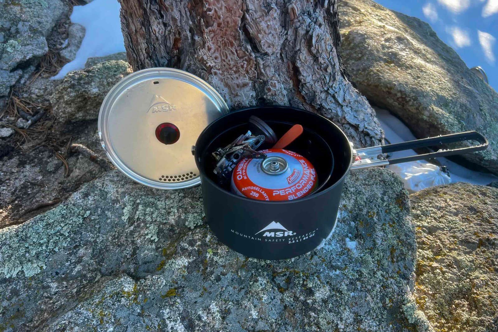 A pot and a lid sits next to a tree. A fuel canister, stove and other accessories are packed inside the pot.