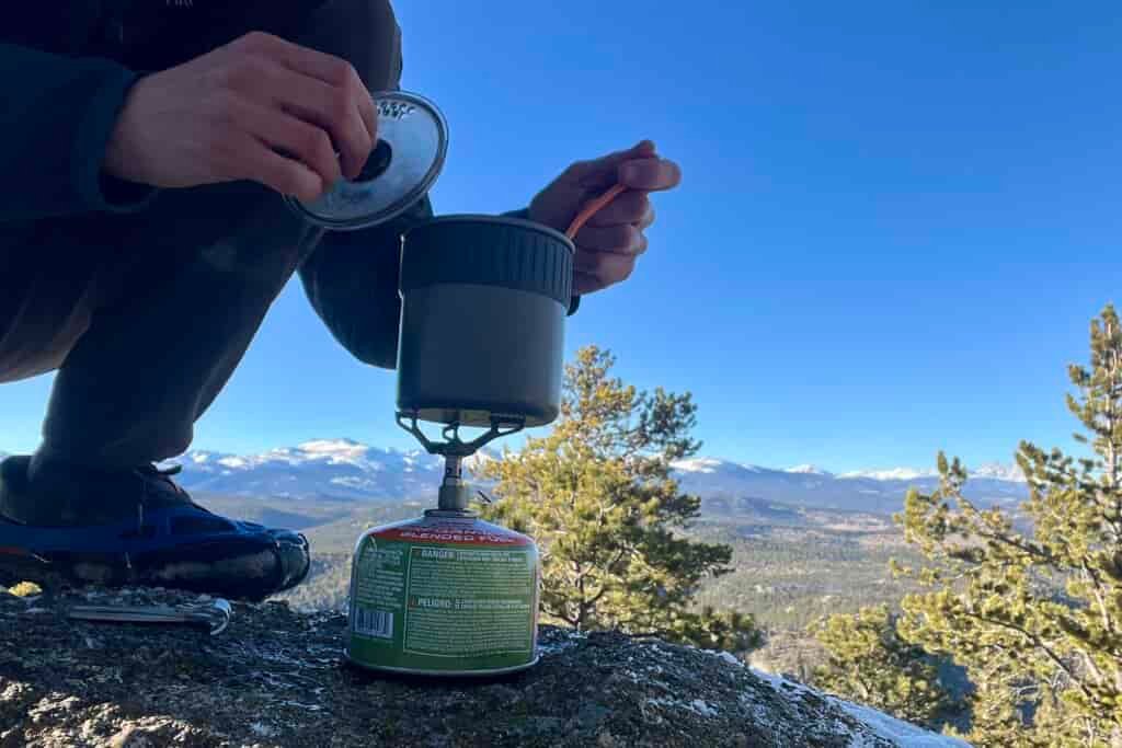 A hiker crouches and cooks with the trail mini solo on a backpacking stove with a mountainous background.