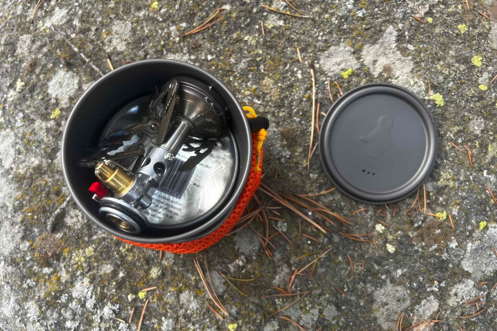 The top down view of a stove and fuel packed into a backpacking pot with the lid next to it sitting on a rock.