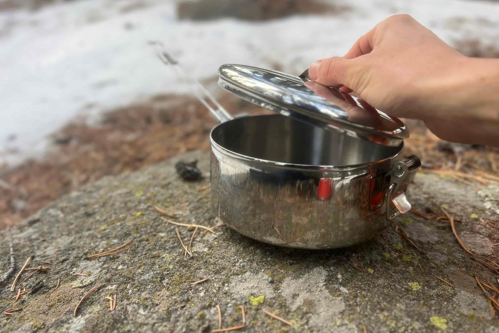 A hand lifts the lid off a steel pot with snow in the background.