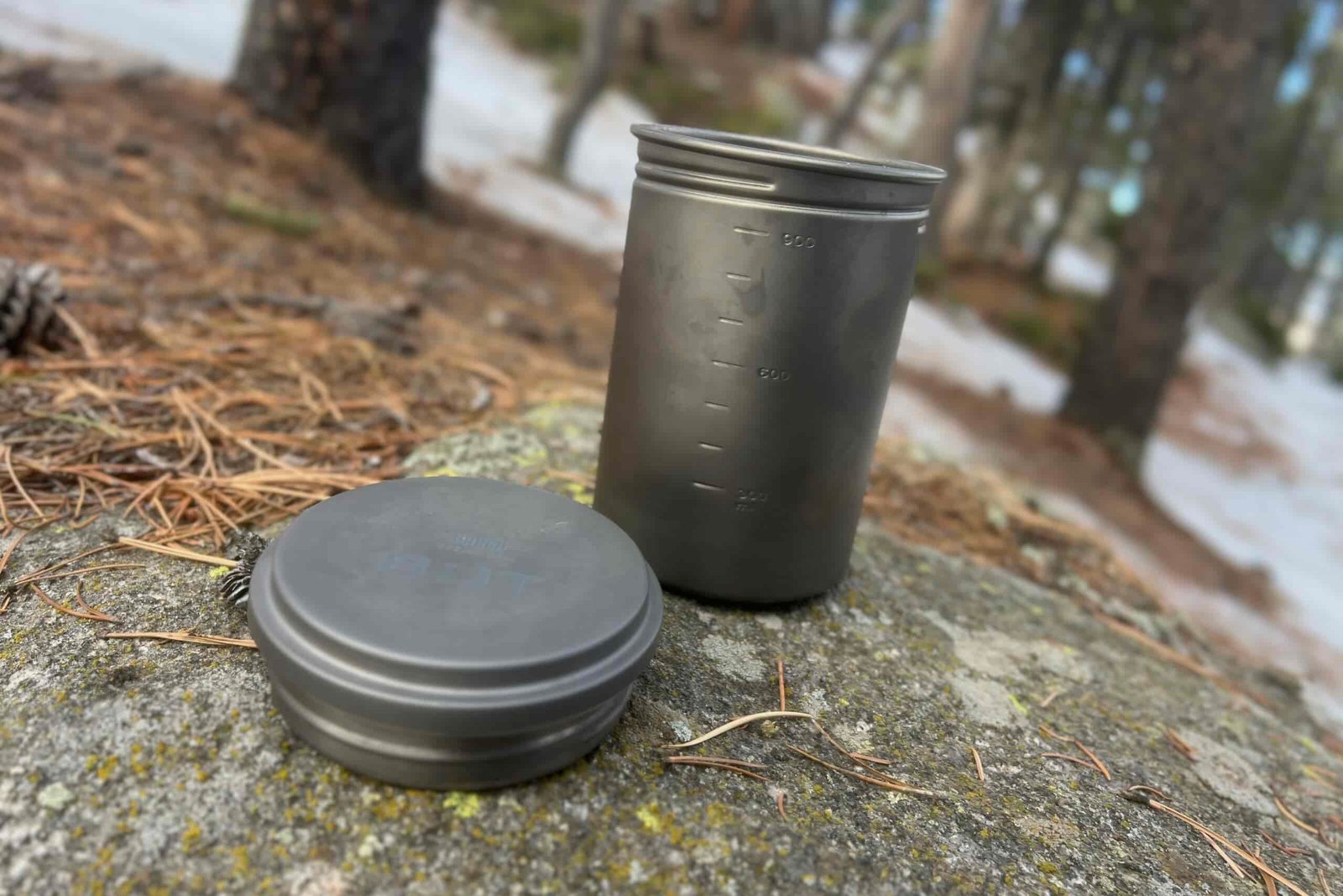 A titanium pot and lid sit on a rock in a forest.