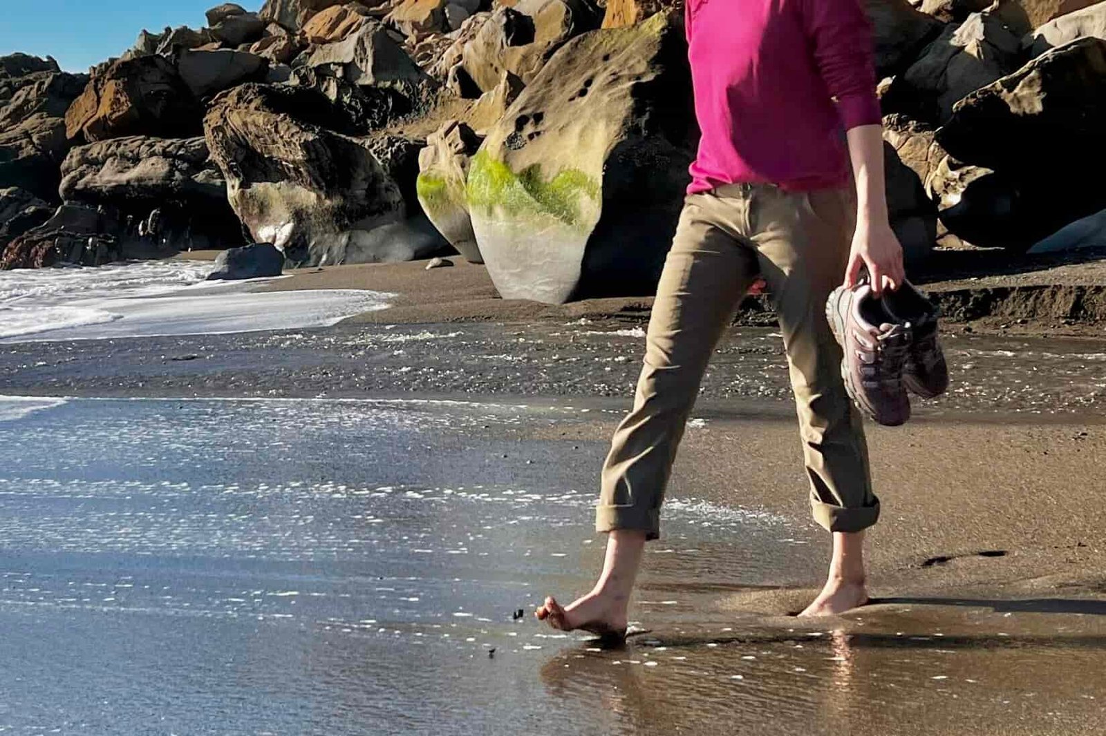 A woman carries the Oboz hiking shoes while walking barefoot on the beach.