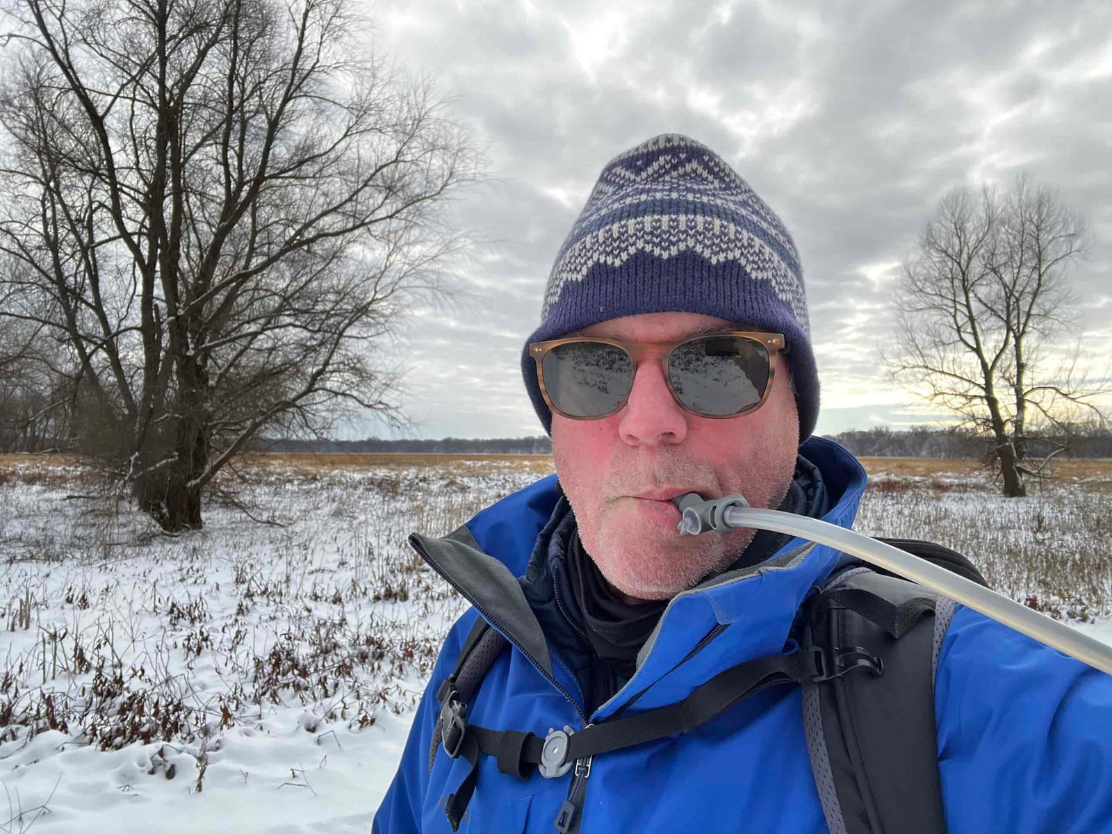 A snowshoer wearing a winter hat and sunglasses drinks from the HydraPak Contour's hydration hose. The background is a snowy field, with an overcast sky.
