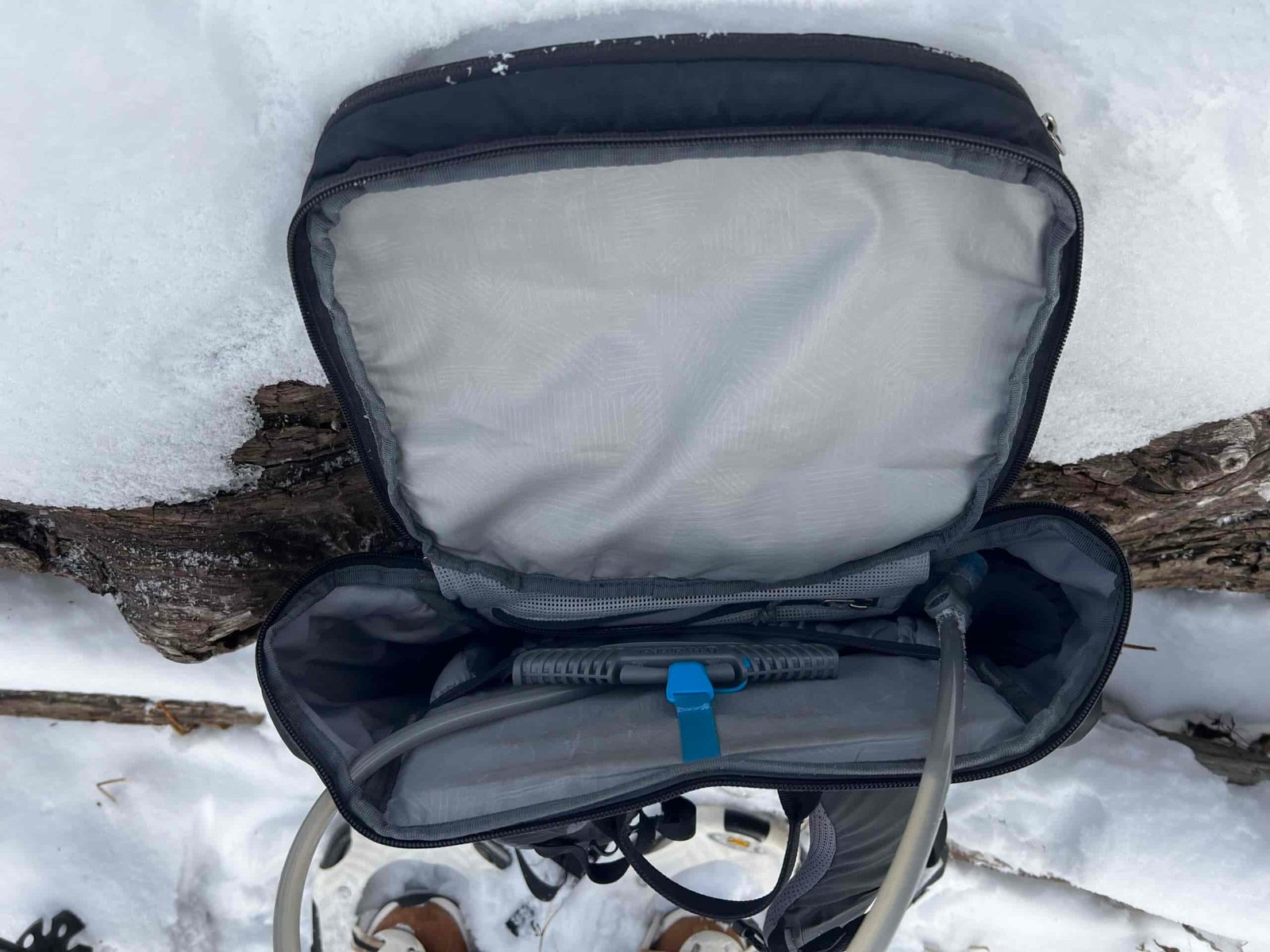 Close up showing the HydraPak Contour loaded into a black backpack. The backpack has a hydration sleeve, a suspension strap that is attached to the Contour, and a hole for the hydration hose to run through. The backpack is sitting on a snowy log.