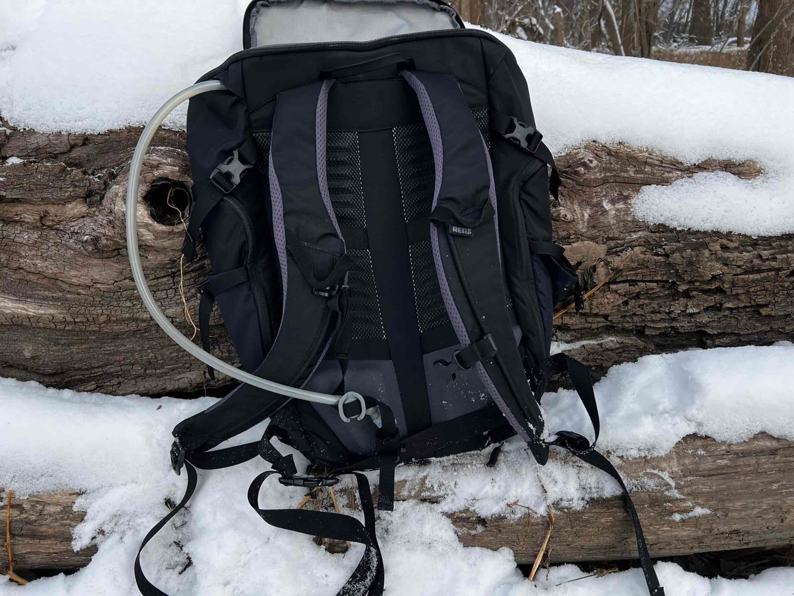 Close-up of a black backpack sitting on a snowy log. The HydraPak Contour's hydration hose is coming out of the top left side of the backpack. It is attached to the sternum strap with a magnet.