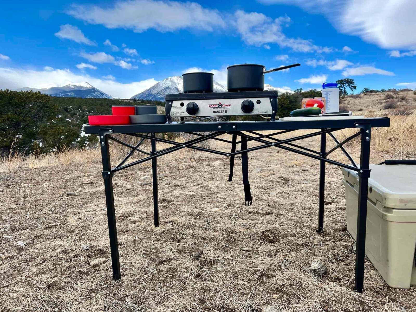 The Mountain Summit Gear Heavy Duty Roll Top Table stands on scrub ground with a scenic mountain view in the background. A two-burner stove, red and gray cookware, and some food items are on top. A beige cooler is sitting on the right side.