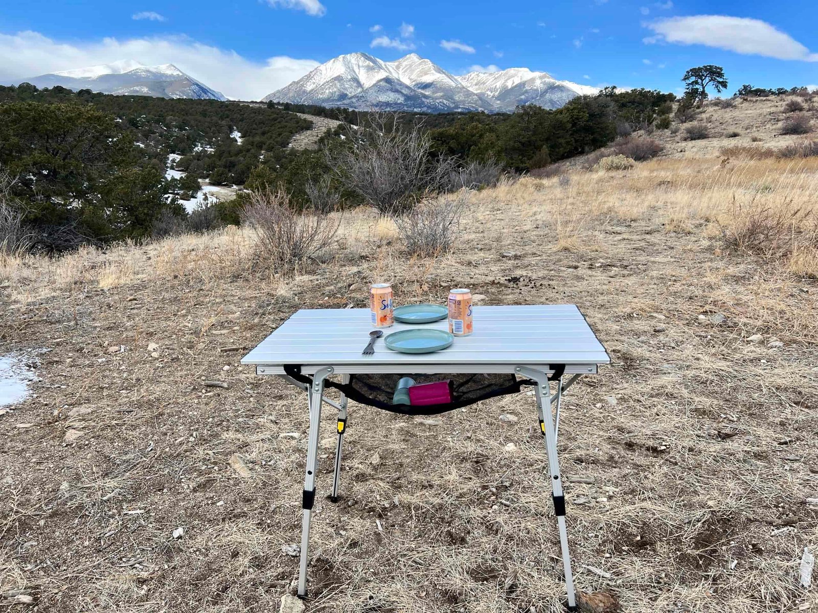 The Portal Quick Adjust Lightweight Roll-Up Table Roll-Top Table stands on scrub with a scenic mountain backdrop. Teal plates and cans of water are on the table. There is a pink coffee bag and a teal coffee maker under the table.