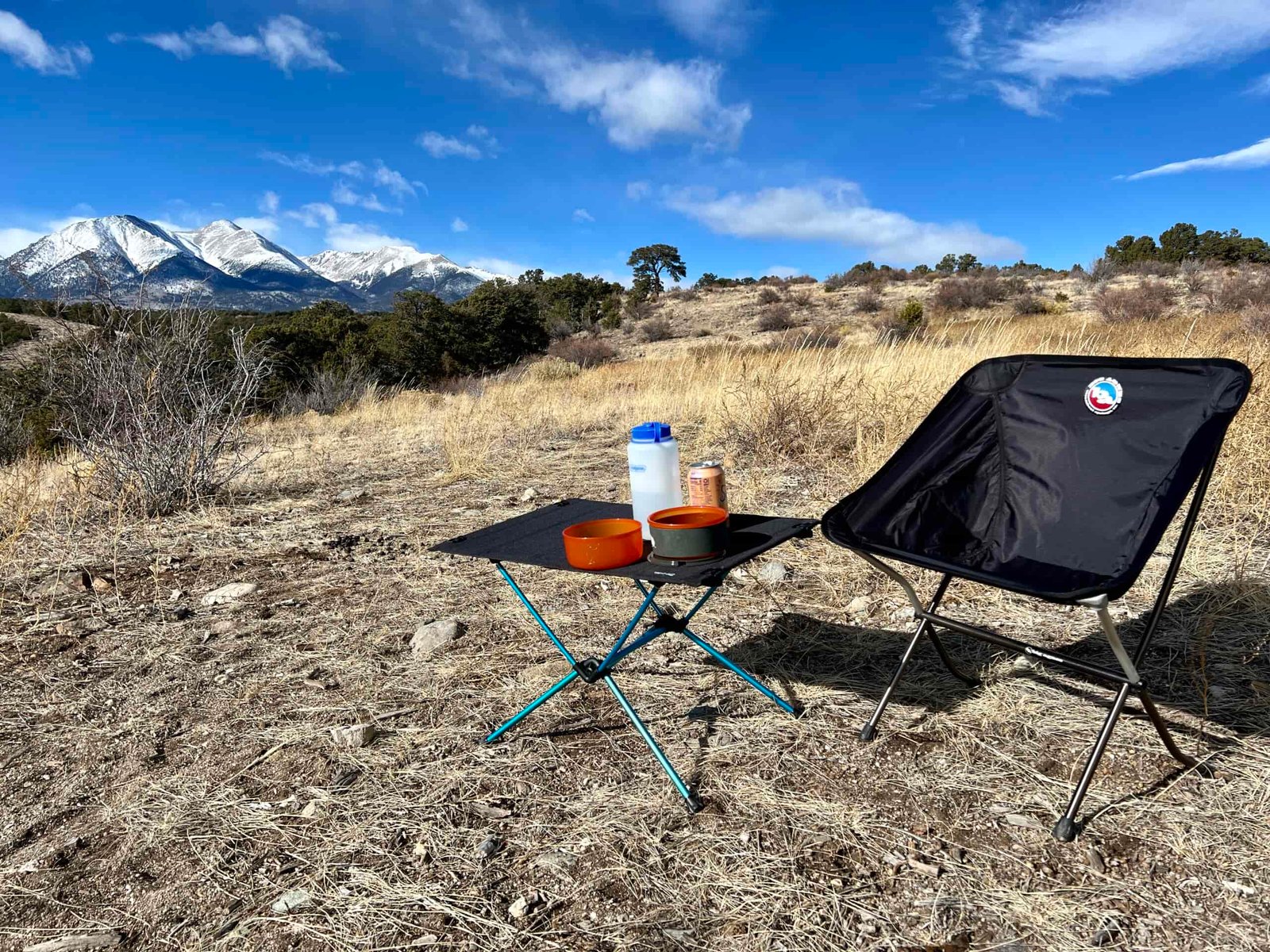 The Helinox One Hard Top Table is standing on dirt and scrub next to an empty black chair. It has orange cookware on it and a Nalgene bottle. The background is a scenic mountainscape.