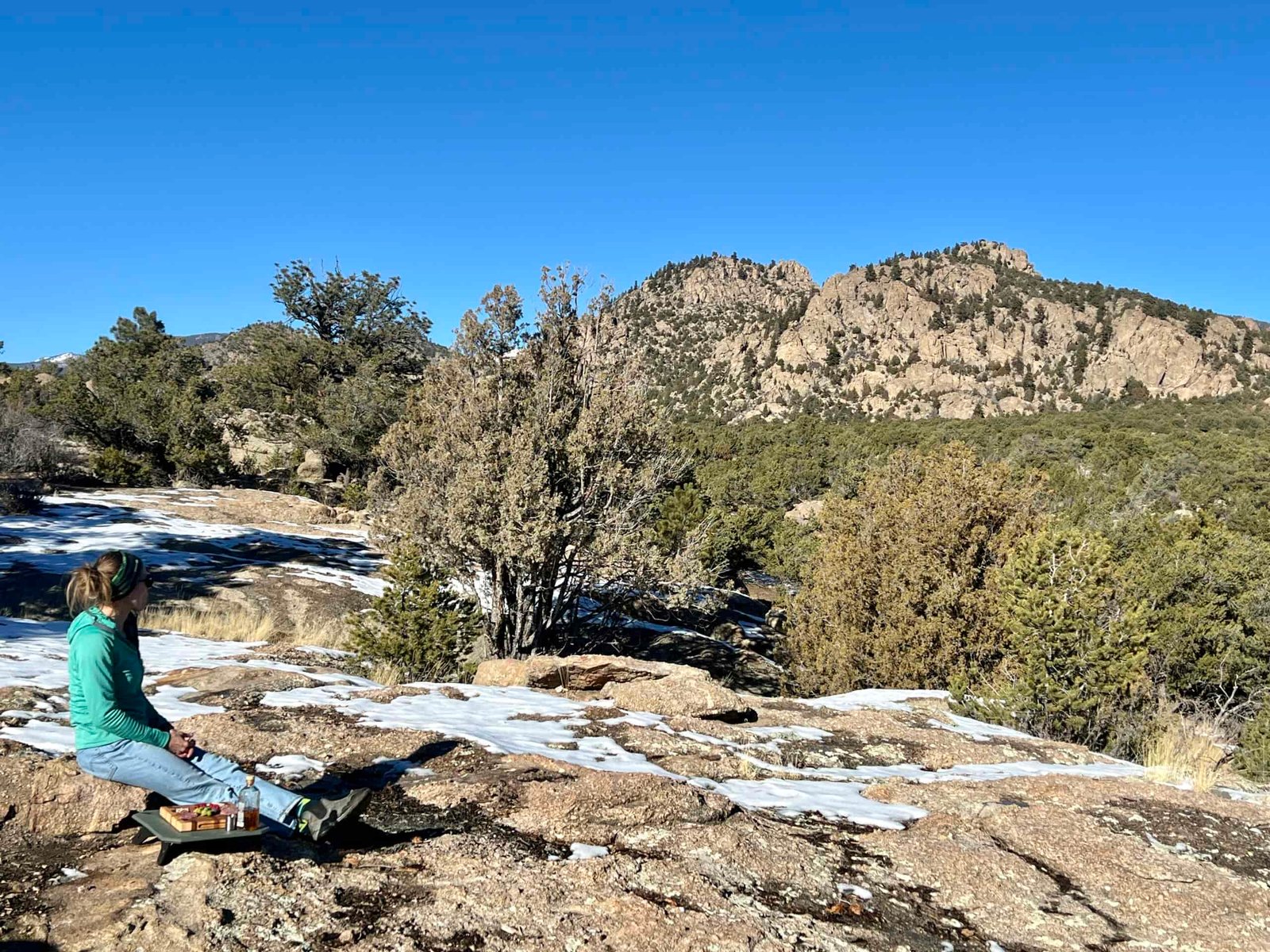 A person sits on a rock looking out into the mountains. Next to her, the NEMO Moonlander Dual-Height Camp Table sits on a rock, with a charcuterie board on top of it.