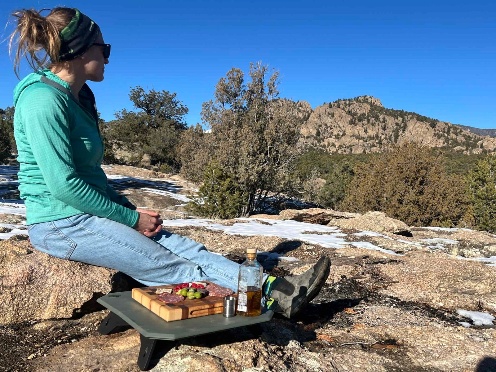 A person sitting on a rock looking out into the distance. The NEMO Moonlander Dual-Height Camp Table is sitting on a rock next to her with a bottle of whiskey and a charcuterie board on top of the table.