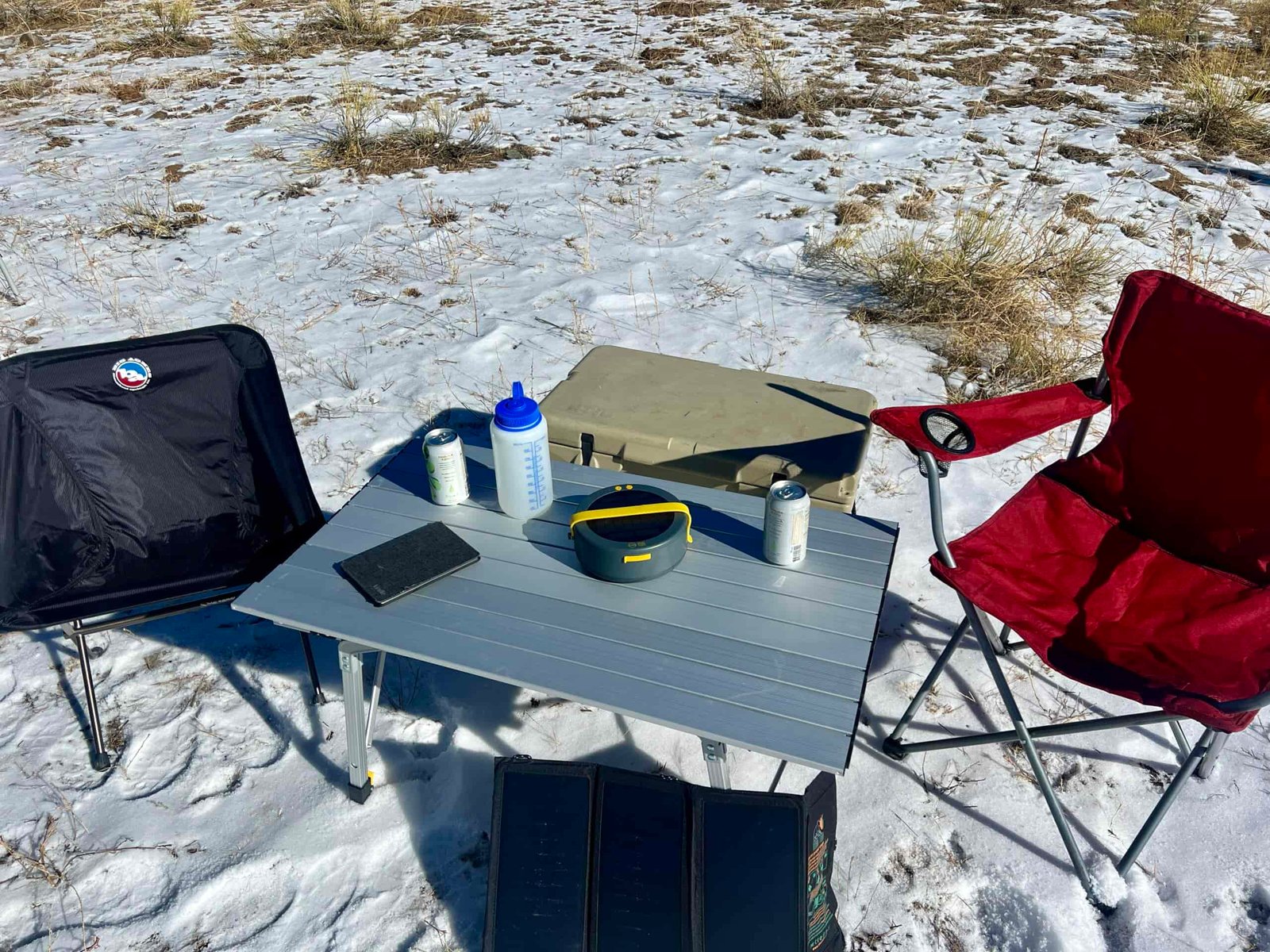 The Portal Quick Adjust Lightweight Roll-Up Table is surrounded by a red chair, black chair, and solar charger. On the top are a Nalgene, kindle, and cans. There is a beige cooler behind the table.