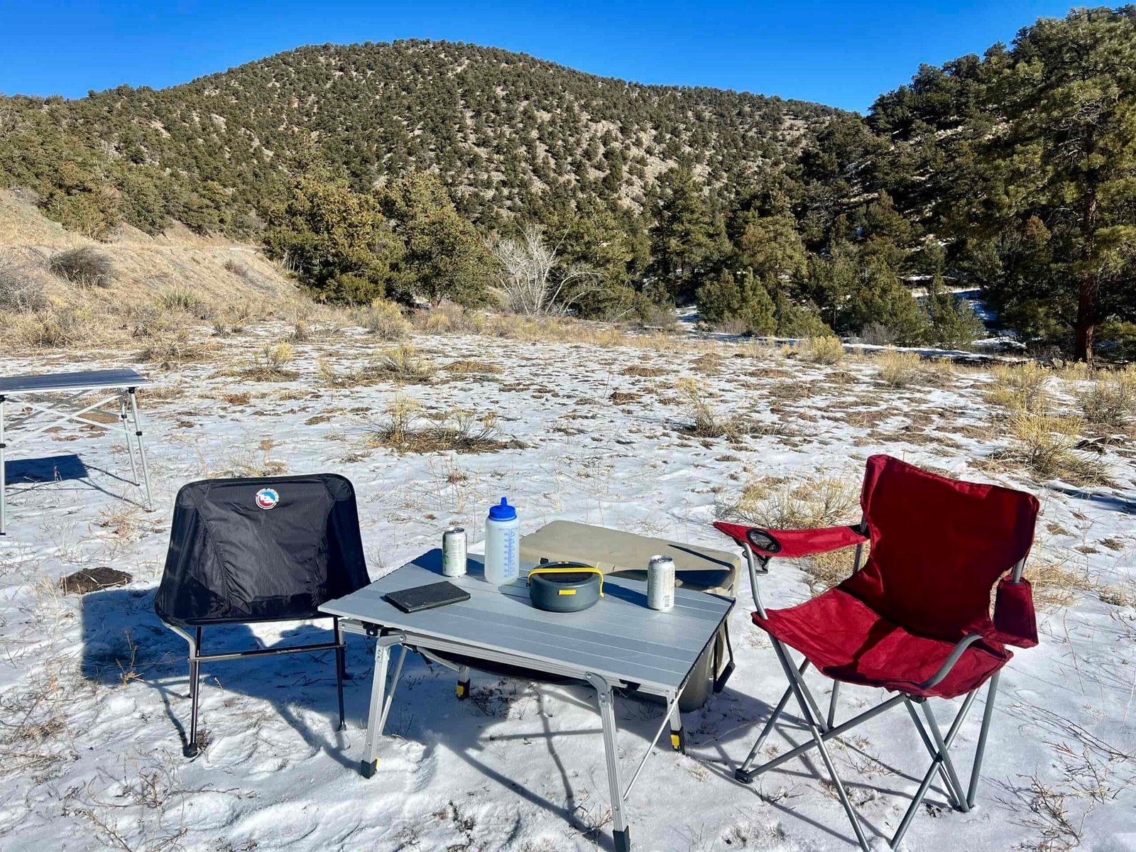 The Portal Quick Adjust Lightweight Roll-Up Table is surrounded by a red chair, black chair, and solar charger. On the top are a Nalgene, kindle and cans. The background