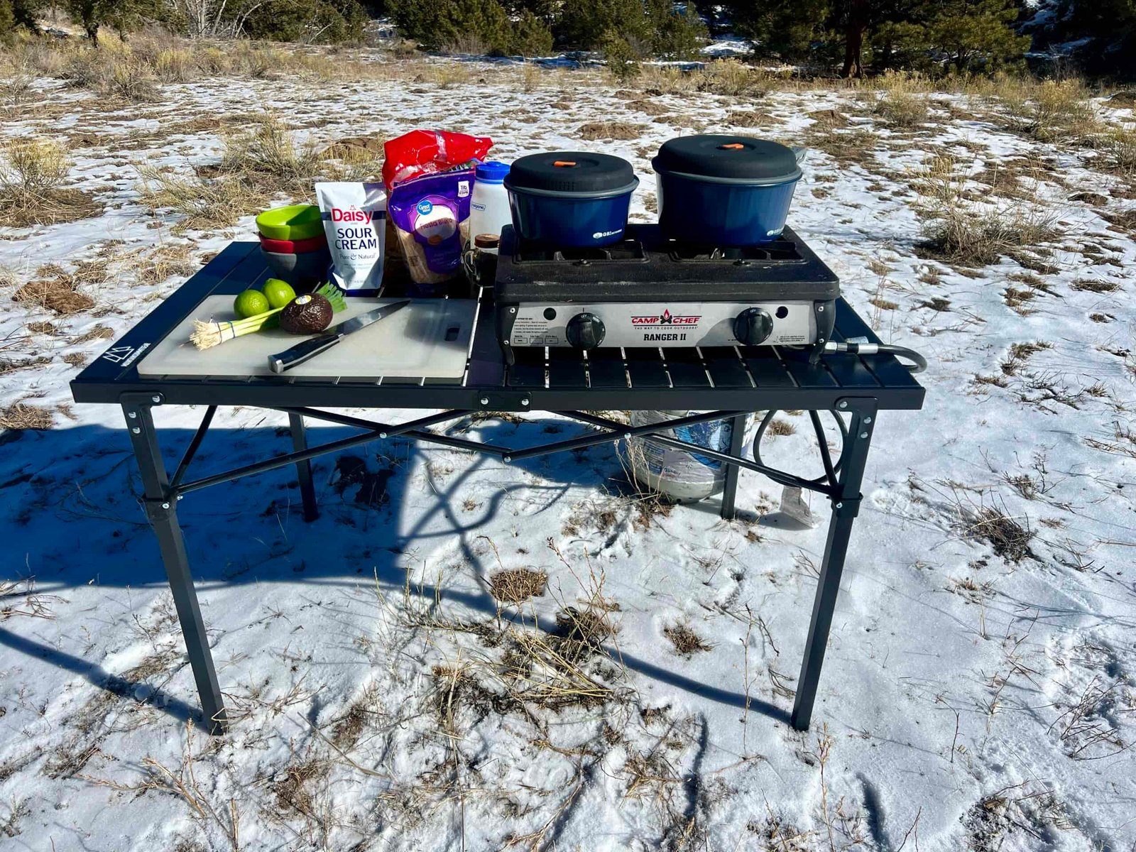 The Mountain Summit Gear Heavy Duty Roll Top Table stands in the snow with tree-covered hills in the background. A two-burner stove and food items are on top. A propane tank sits on the ground behind it.
