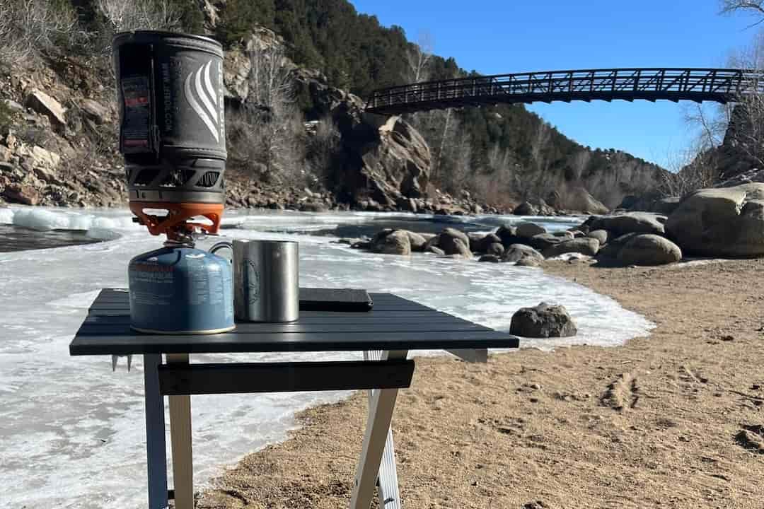 The GCI Compact 20 stands on a sandy river beach. The river is mostly iced over. A Jetboil, stainless cup, and kindle sit on top of the table. In the background, a bridge crosses the river.