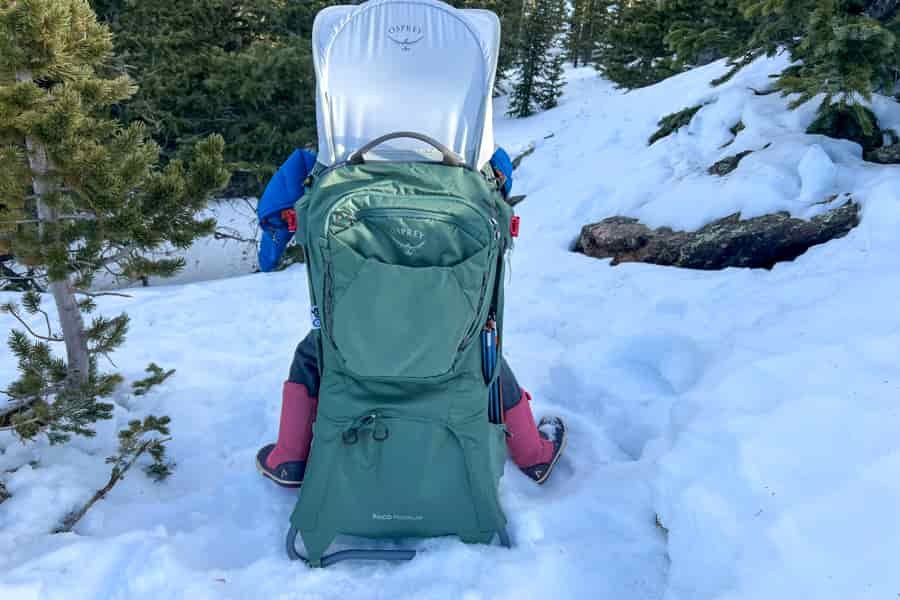 A back view of the Osprey Poco Premium baby carrier on a snowy trail, showcasing its sleek green design and sunshade.