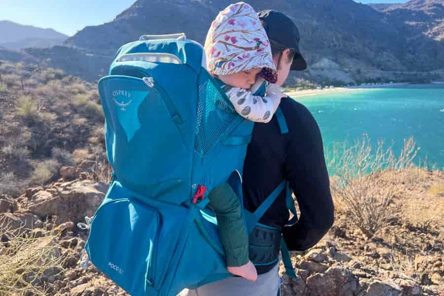 Hiker wearing the Osprey Poco SLT carrier while overlooking a scenic coastal view, with a baby comfortably seated in the carrier.