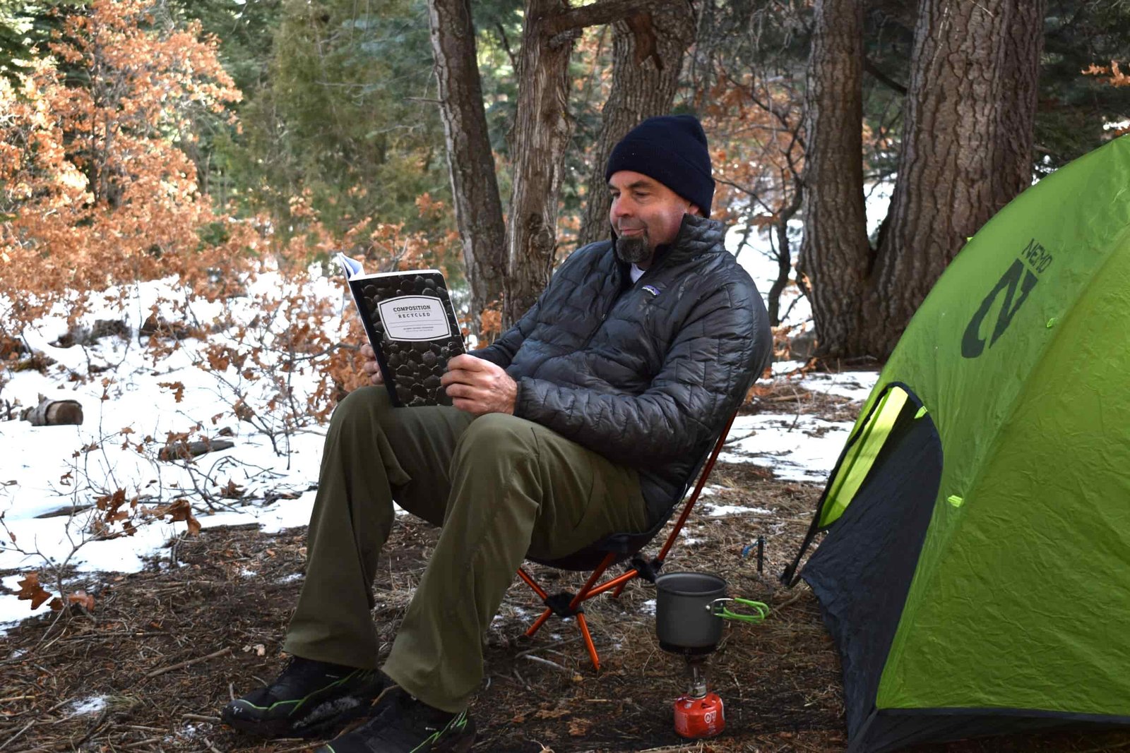 A male backpacker reads a hiking journal while sitting on a backpacking chair next to a tent in the snow