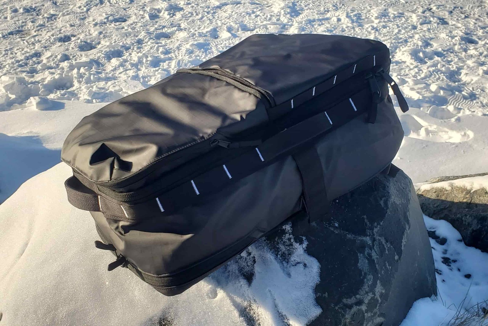 A black backpack sits on a snow covered rock in the sun.