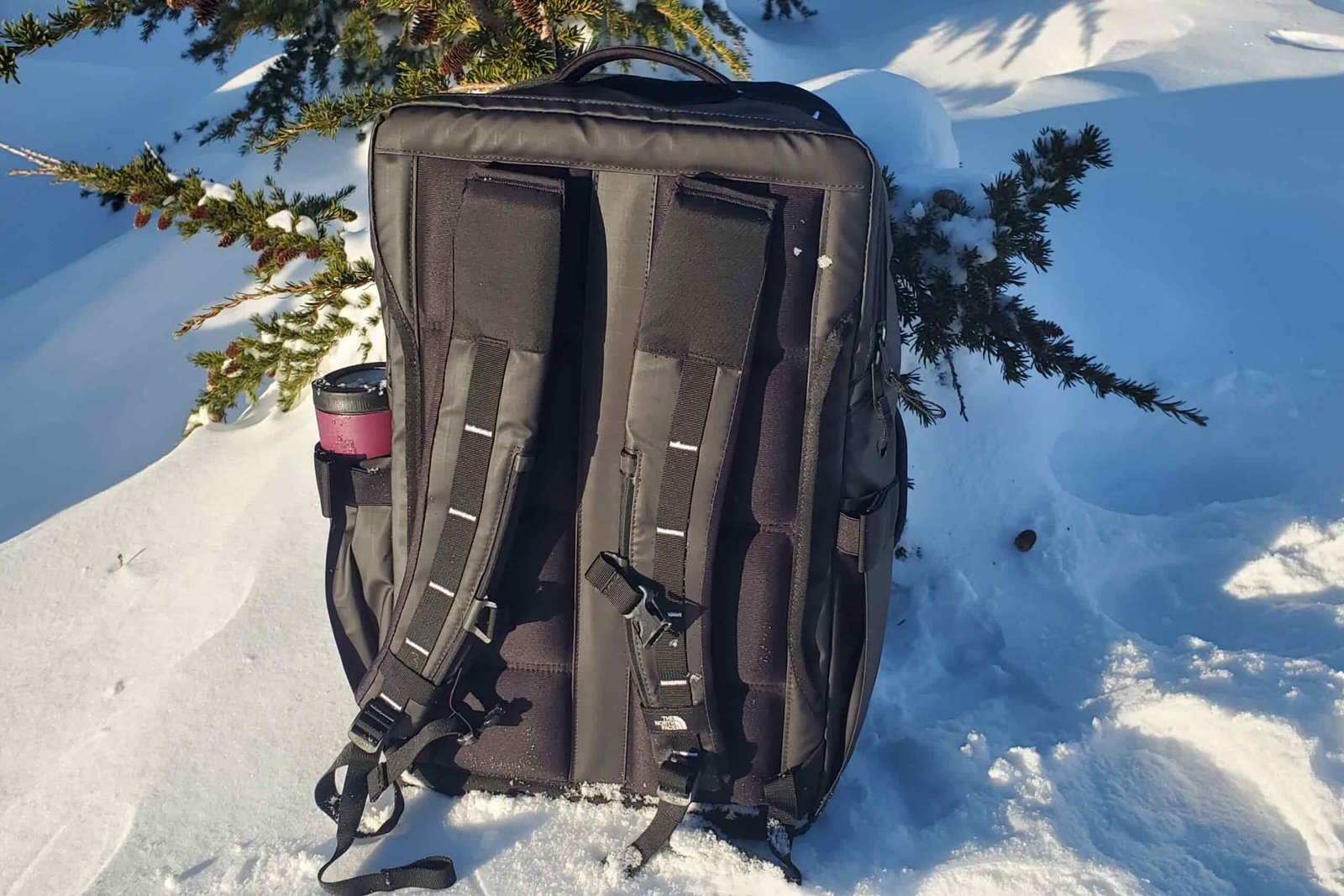 A black backpack sits in the snow in the sun.