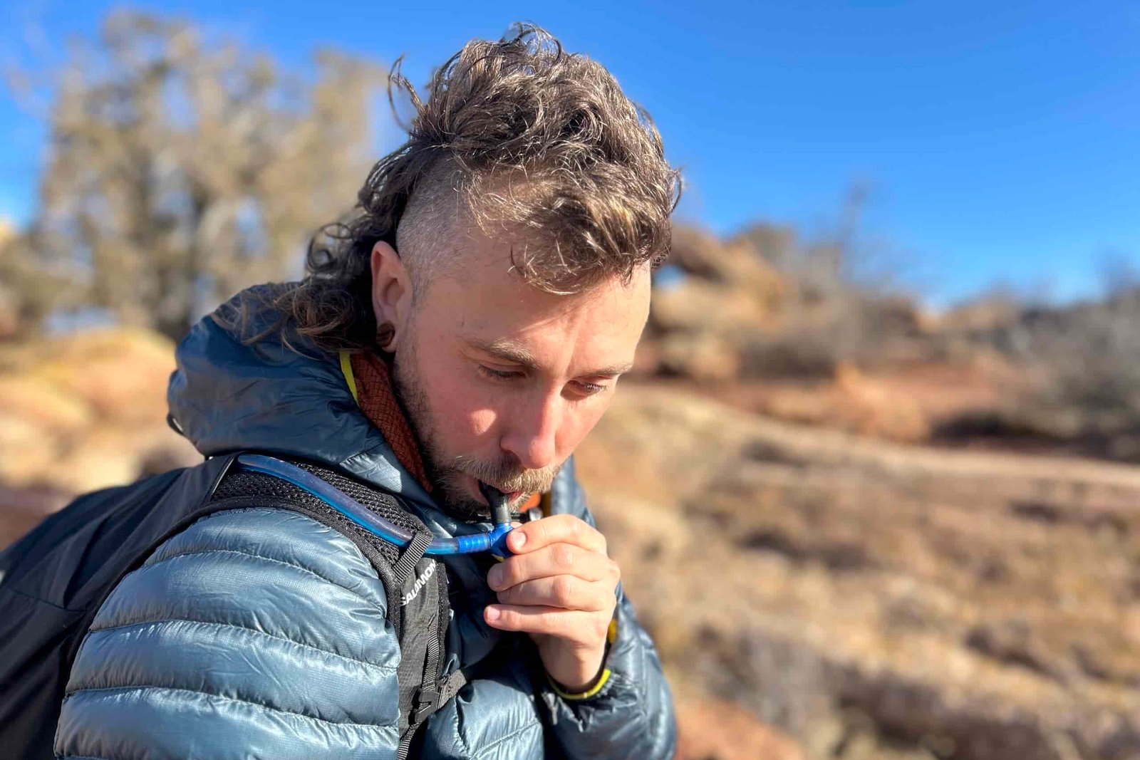 Close-up image of a man drinking water out of a hydration tube routed along his shoulder strap.