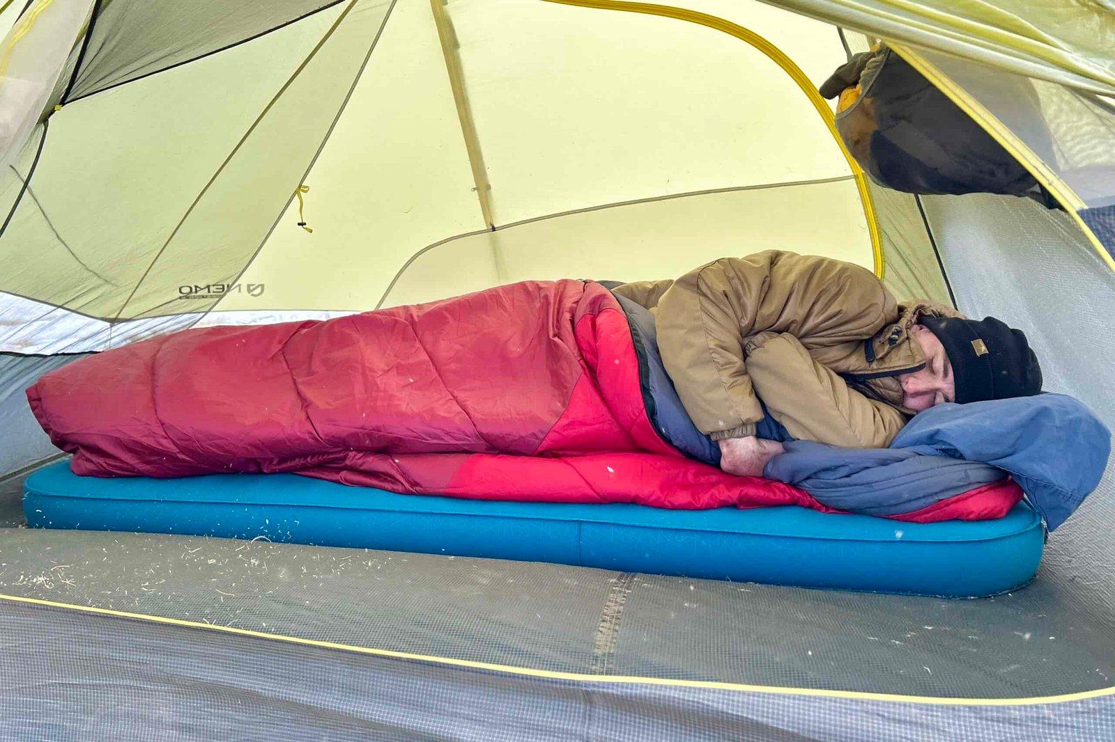 A man in a jacket and beanie sleeps solo in a thick sleeping bag inside of a tent.