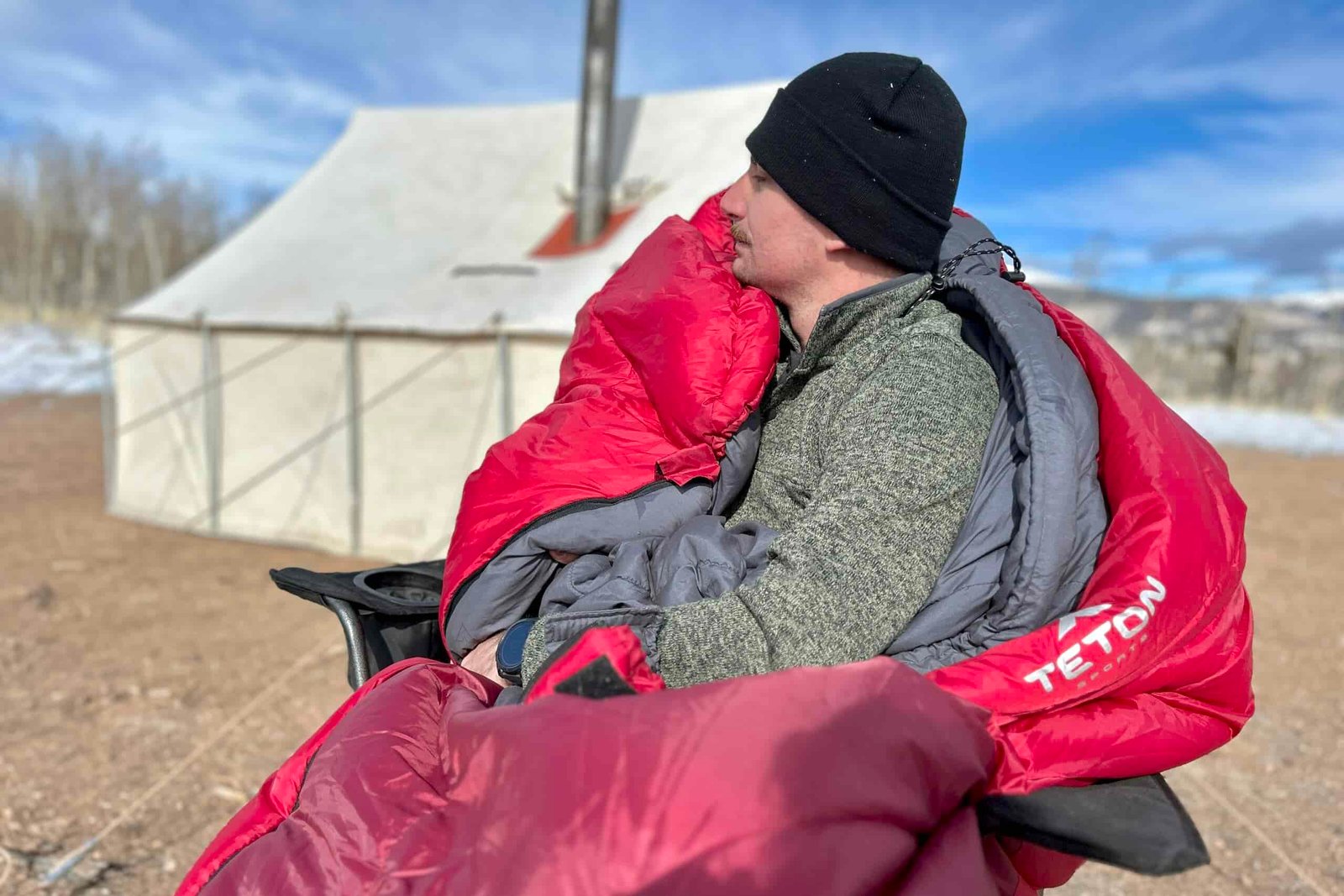 A man sits in a chair in a big, cushy sleeping bag with a canvas tent and snowy mountains in the background.