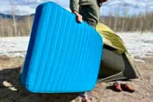Close-up image of a person loading a wide sleeping mattress into a tent in a snowy landscape.