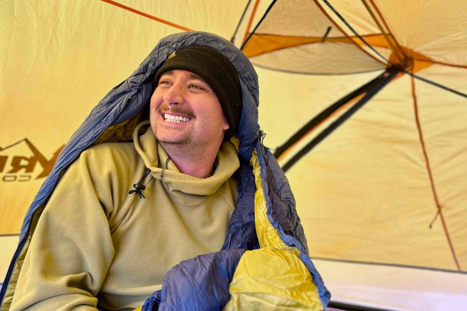 A man smiles off to the side inside of a tent. His sleeping bag hood is over his head.