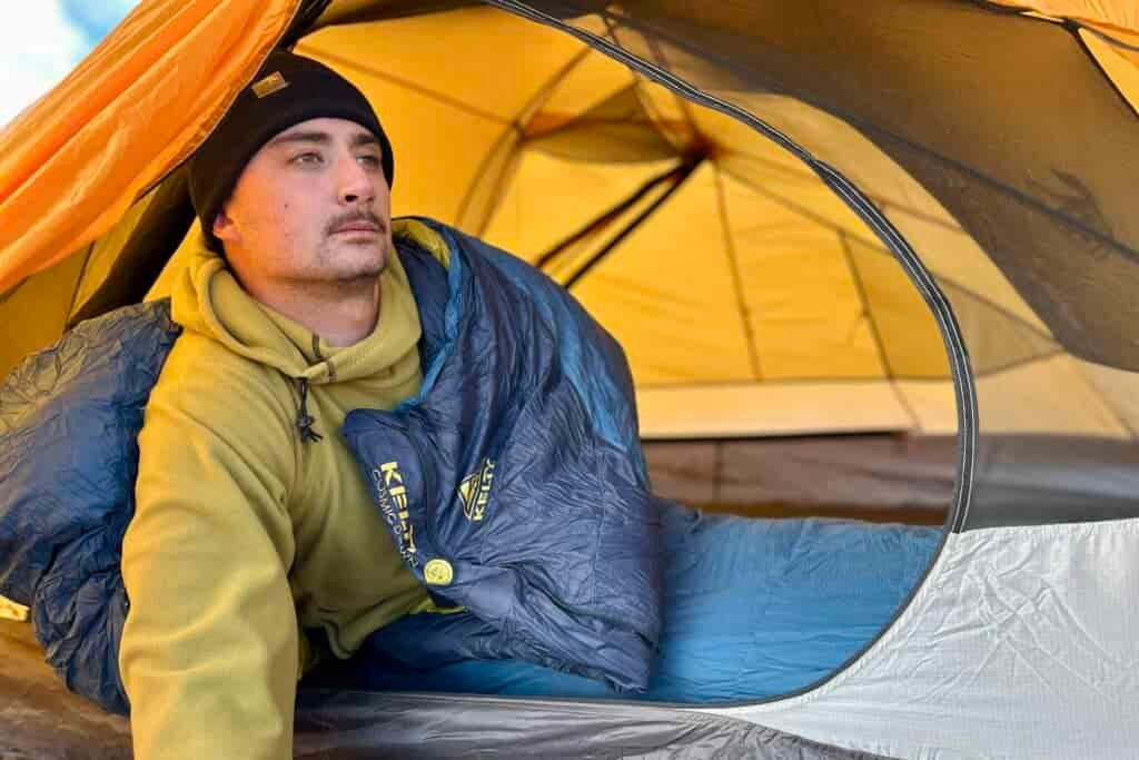 A man looks out of a tent to take in the view while still in his sleeping bag.