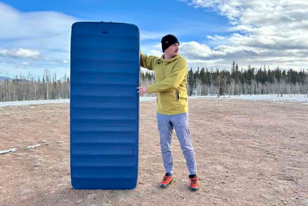 A man in a snowy, forested landscape stands next to a tall sleeping pad.
