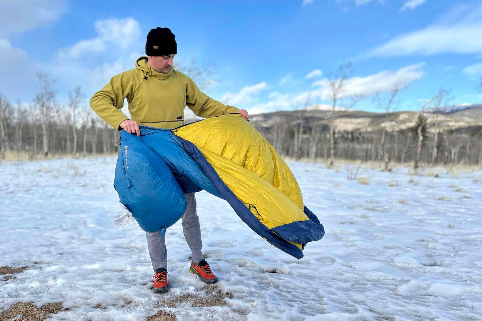 A man fluffs a down sleeping bag outside in a snowy mountain landscape.
