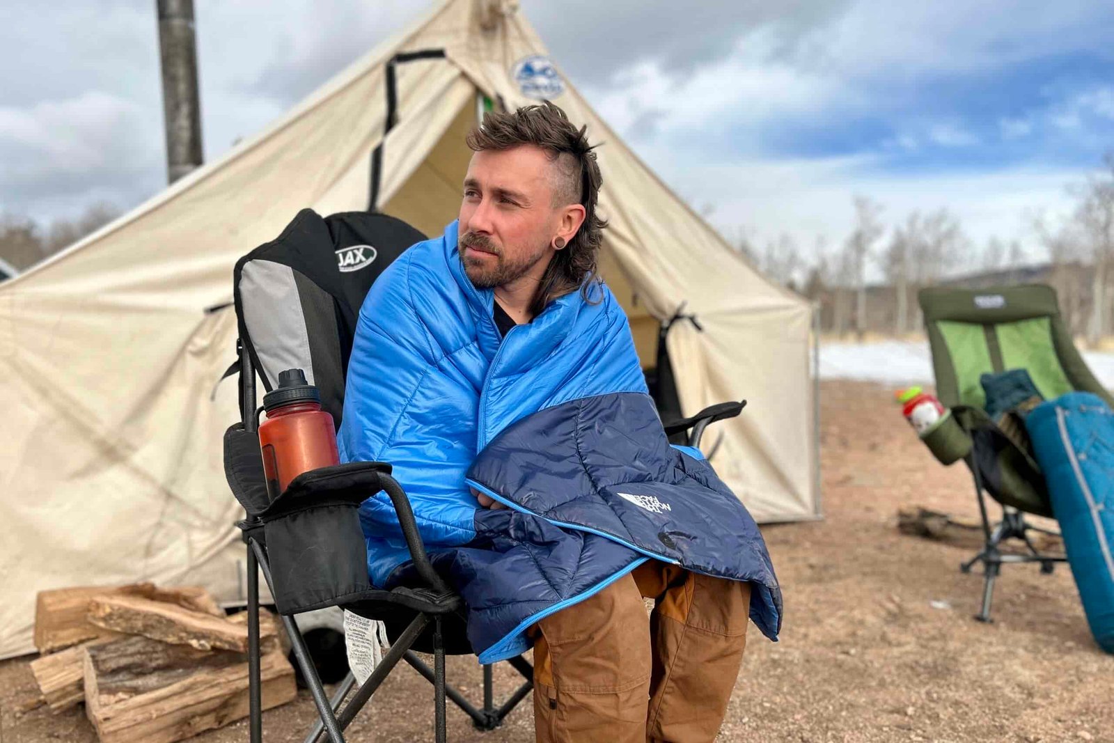 A man sits in a chair in front of a large canvas tent. He is wrapped in a down blanket and there's snow in the background.