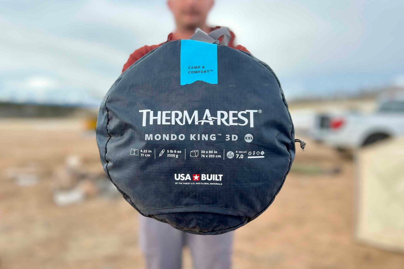 A person holds up a sleeping pad in a package to show off the bottom, where the text reads "THERM-A-REST MONDO KING 3D".