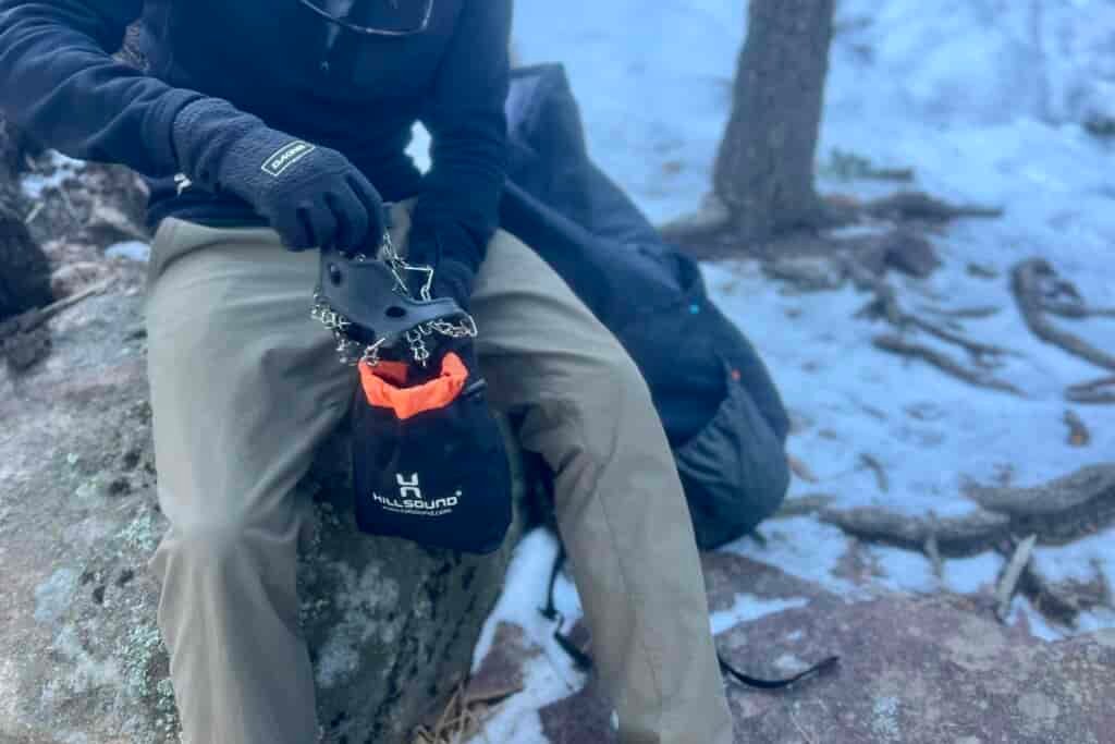 A hiker sits on a rock and packs ice spikes into a stuff sack against a snowy forest background.