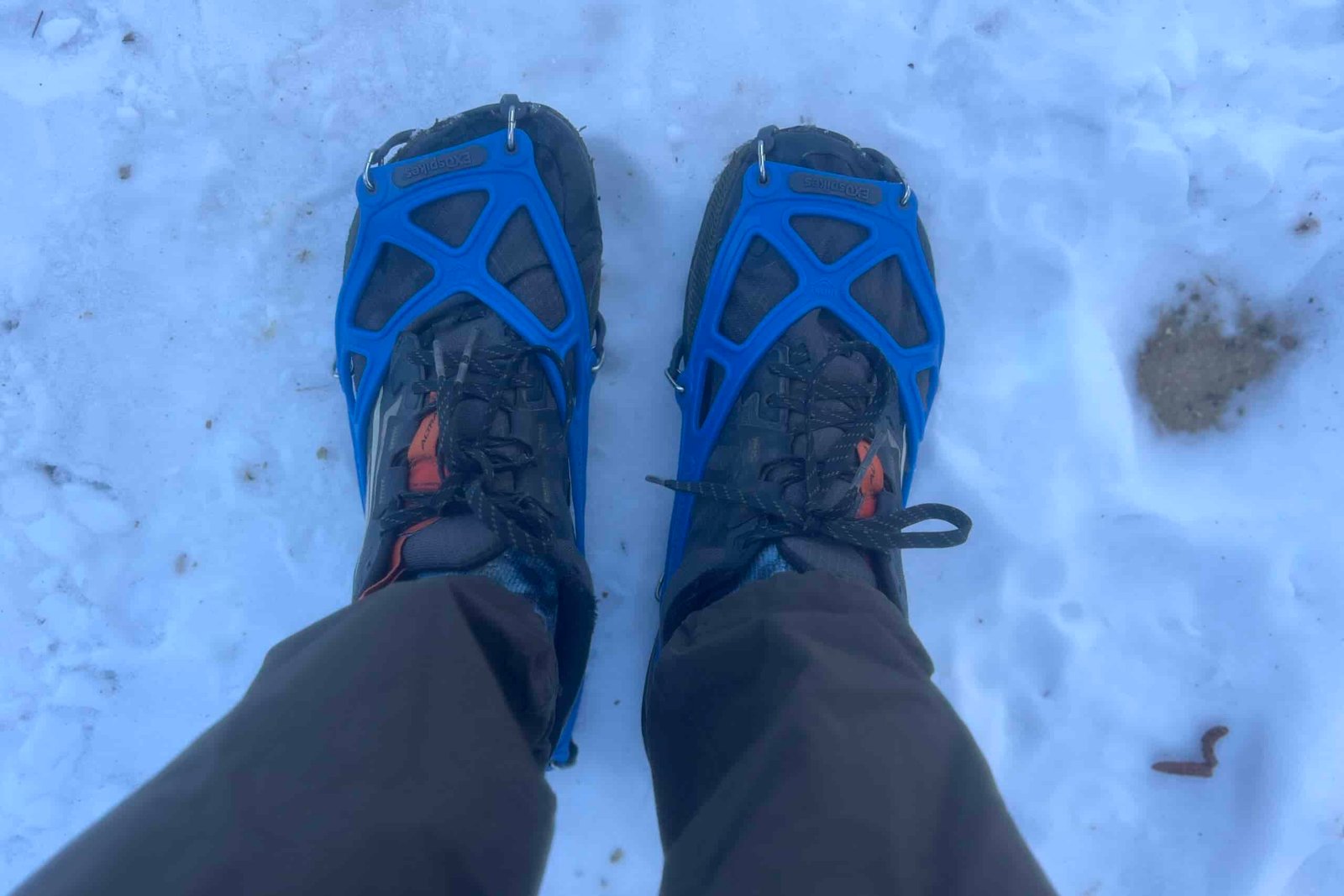 Top down view of a pair of feet standing on snow while wearing trail runners and ice cleats.
