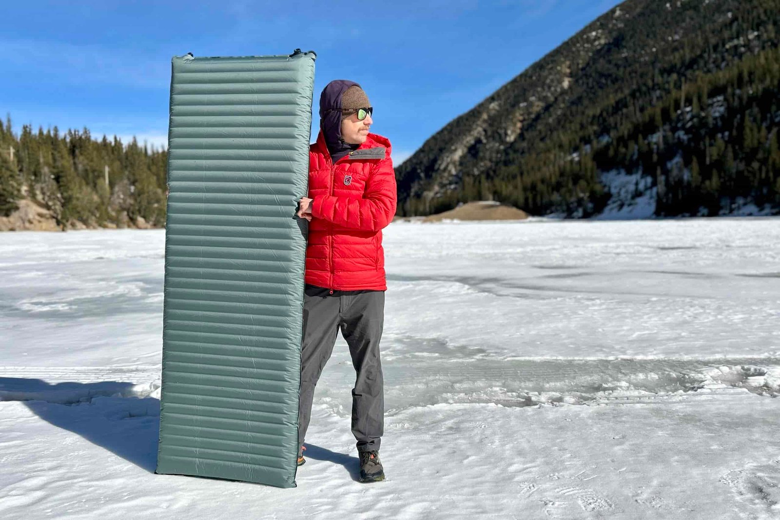 A man holds a camping mattress vertically in a snowy mountain setting.