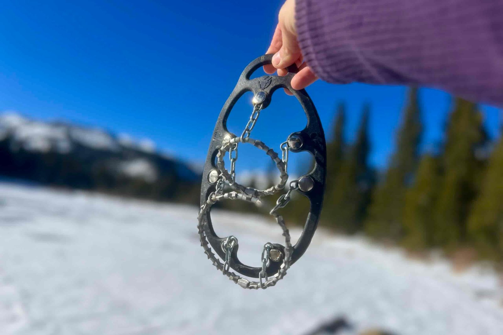 A hand holding up a pair of ice cleats against a wintery mountain background.