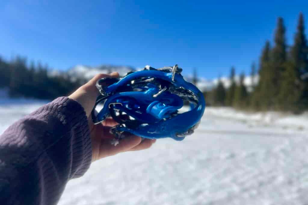 A hand holds a pair of ice cleats folded in half with a snow mountain background