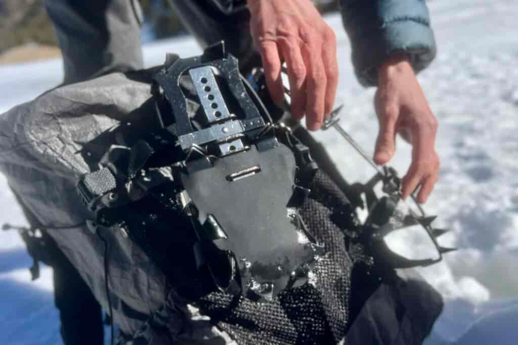 A hiker's hands hang a pair of trail crampons on a backpack with snow in the background.