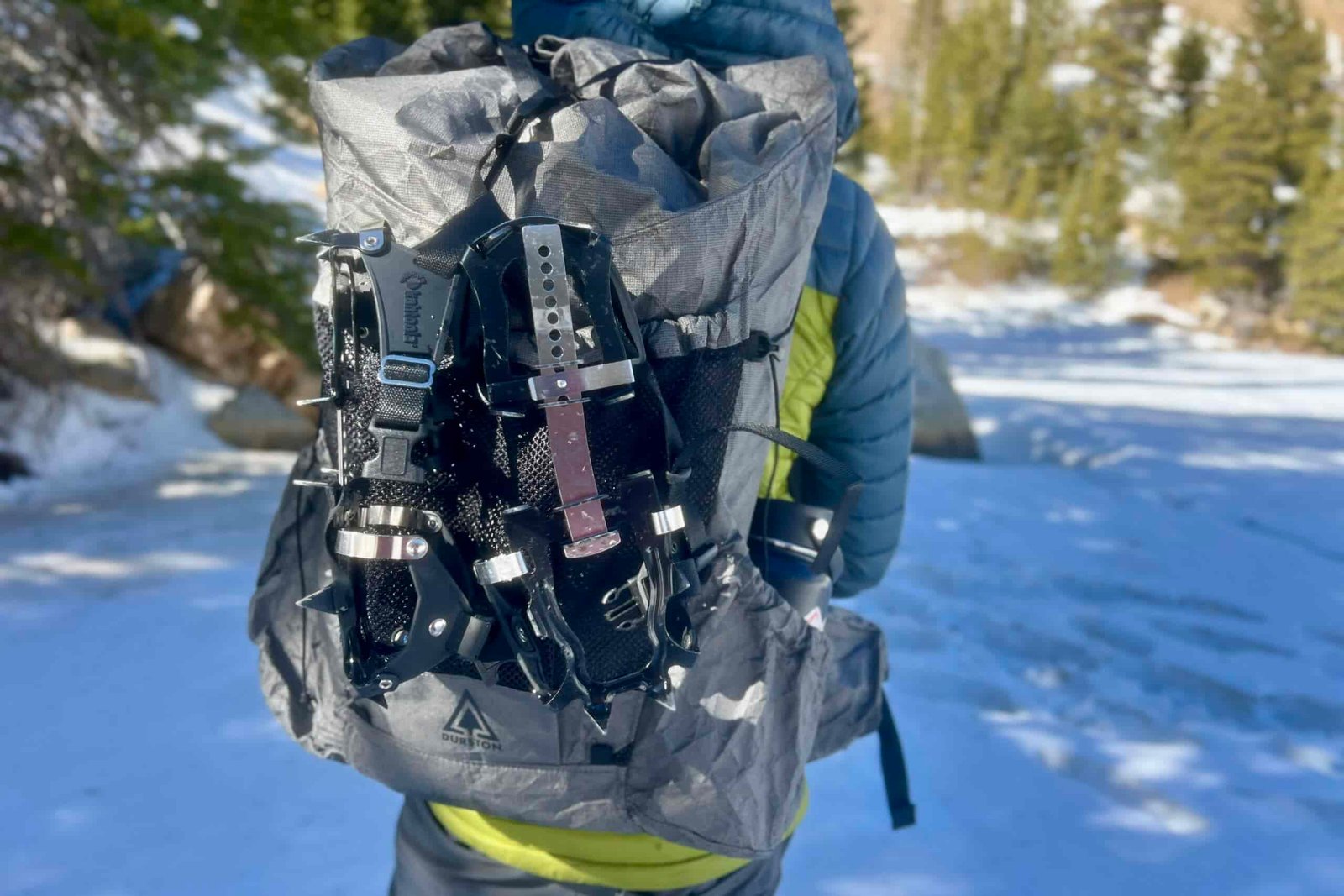A hiker facing away from the camera wearing a backpack with trail crampons strapped to the back. Snow and trees in the background.