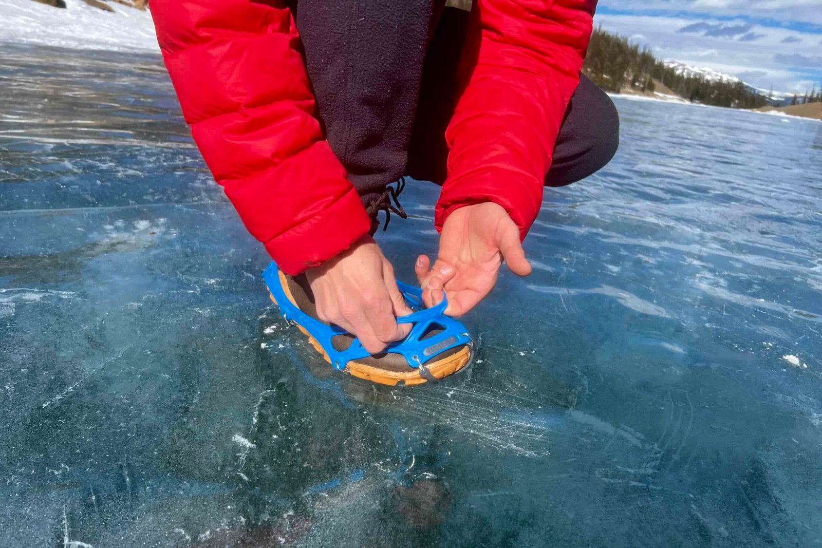 A hiker kneels on a frozen lake and adjusts his ice cleats on his feet.