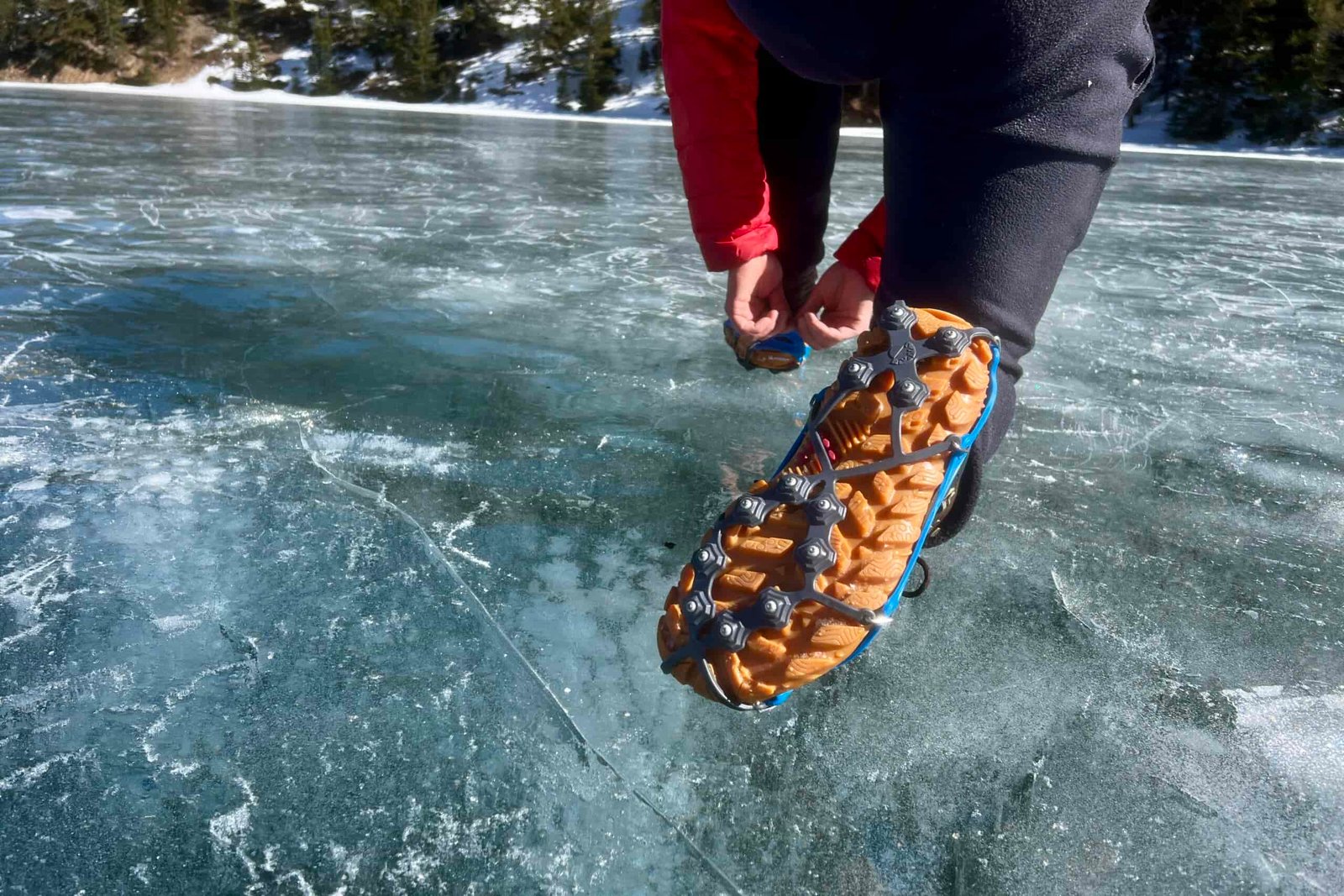 A hiker kneels on an icy lake showing the bottom of his boot wearing ice cleats.