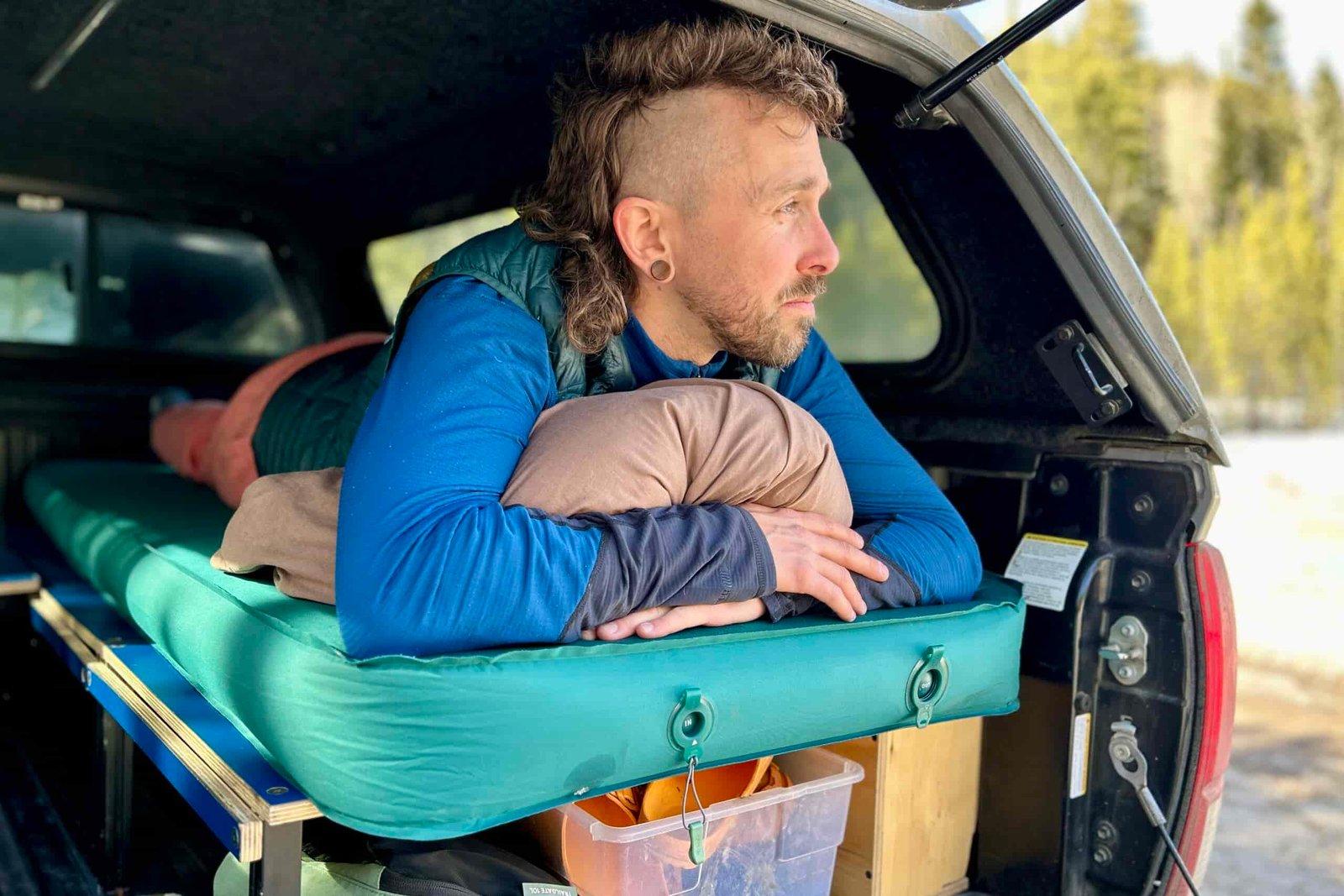 A man rests on a camping mattress in the back of a truck, looking out of the back.