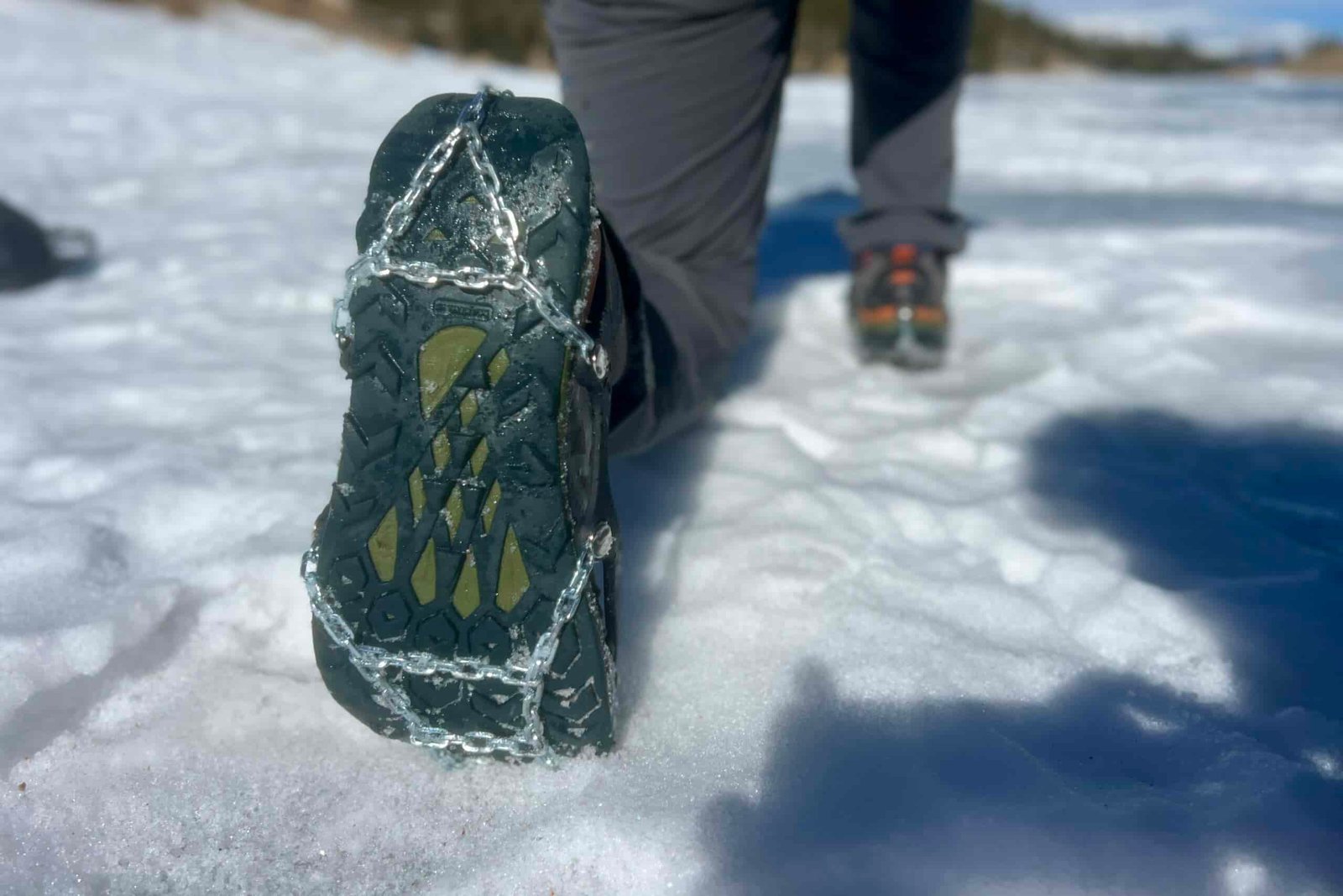 A hiker kneels on snow, showing the bottom of his shoe which is wearing an ice cleat.