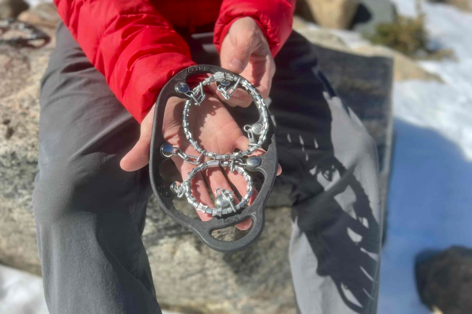 A hiker sits on a rock and uses his hands to display a pair of ice cleats.