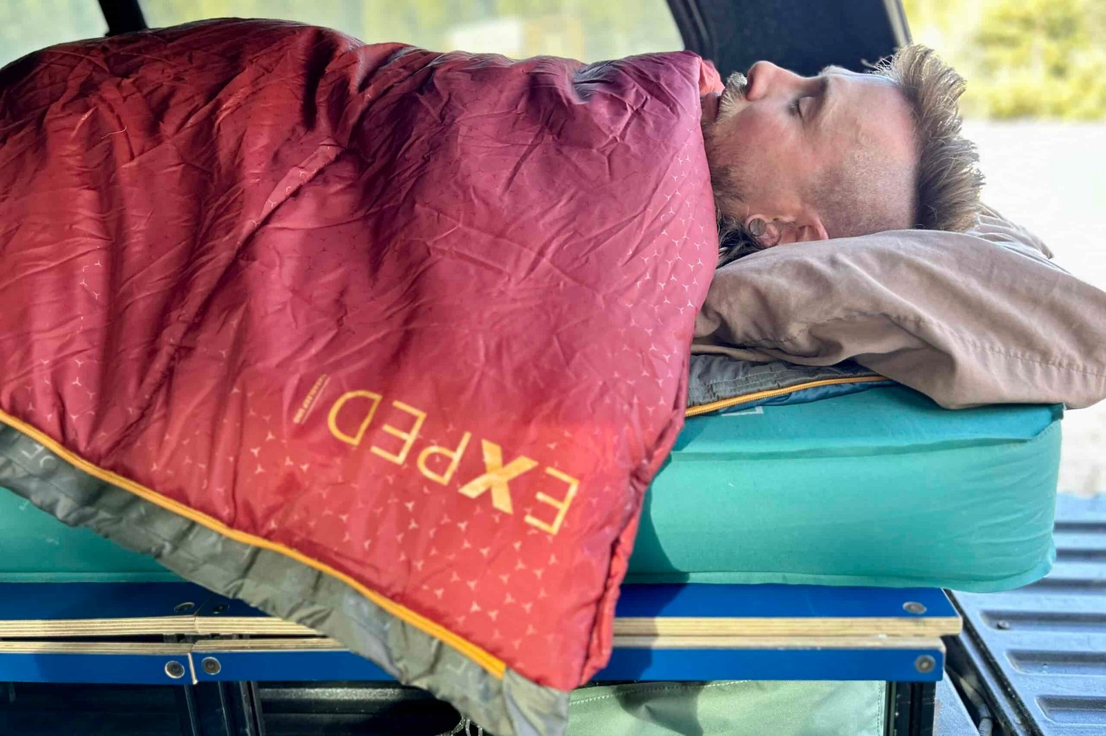 Side-profile of a man resting on a camping mattress in the back of a truck with a sleeping bag pulled to his chin.