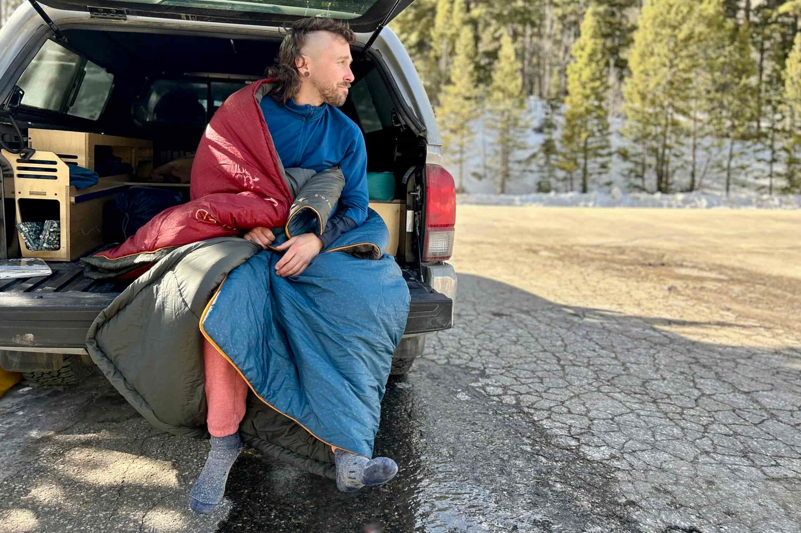A man sitting on the tailgate of a truck with a sleeping bag wrapped around him like a blanket.